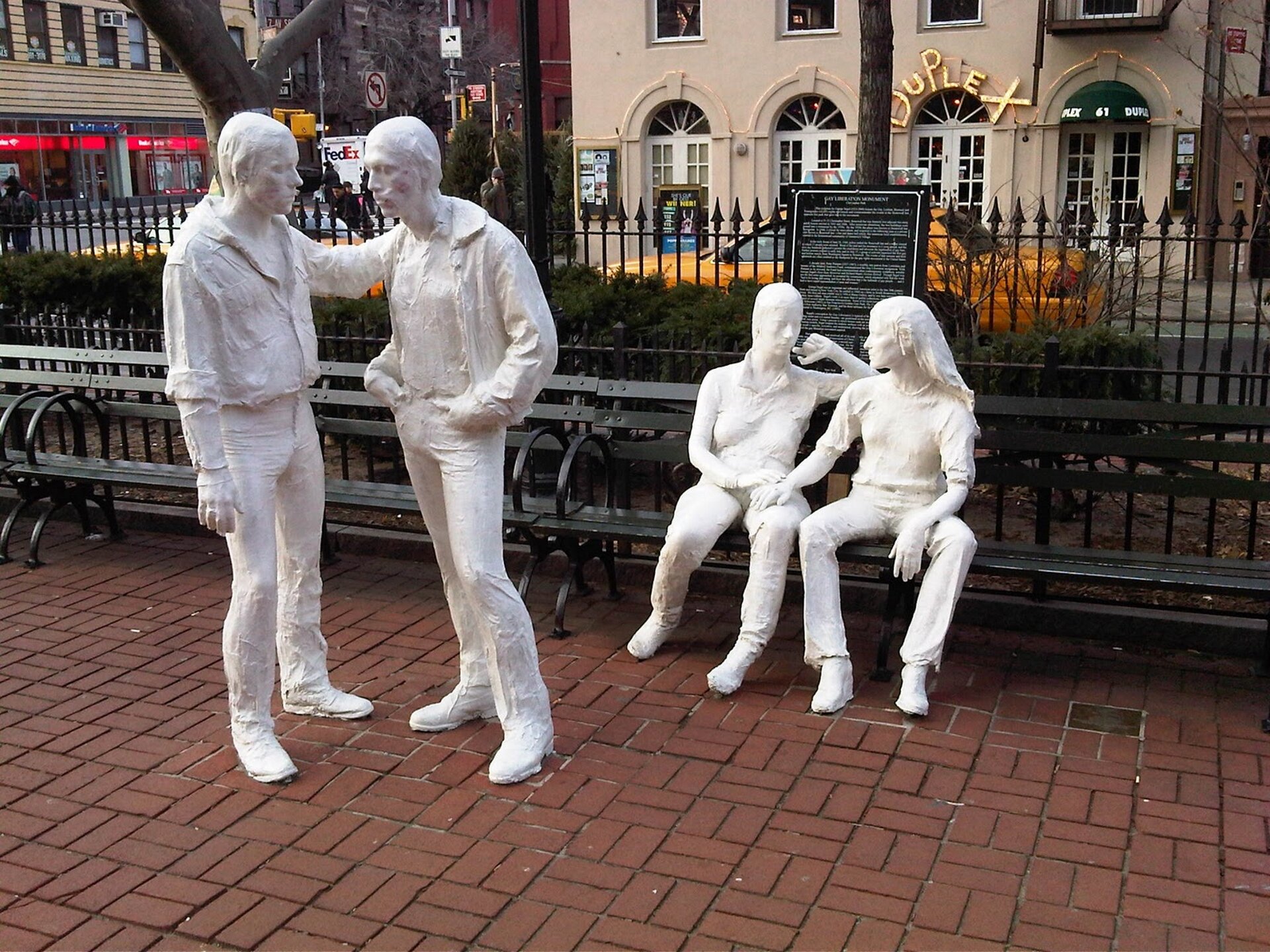 Fotografia przedstawia pracę „Gay Liberation”, autorstwa George Segala.  Rzeźba ta składa się z czterech figur – dwóch stojących mężczyzn i dwóch kobiet siedzących na ławce. Figury zostały wykonane z brązu i pomalowane na biało, są naturalnej wysokości. Na pierwszym planie znajdują się dwaj mężczyźni , jeden obejmuje drugiego za ramię, obie figury „spoglądają” na siebie, jedna z figur (ta z prawej strony) ręce trzyma w kieszeniach spodni. Tuż za nimi widoczne są dwie kobiety – kobieta z prawej strony trzyma rękę na udzie drugiej kobiety, obie figury czule patrzą sobie w W tle fotografii widoczne są charakterystyczne żółte taksówki oraz budynki. 