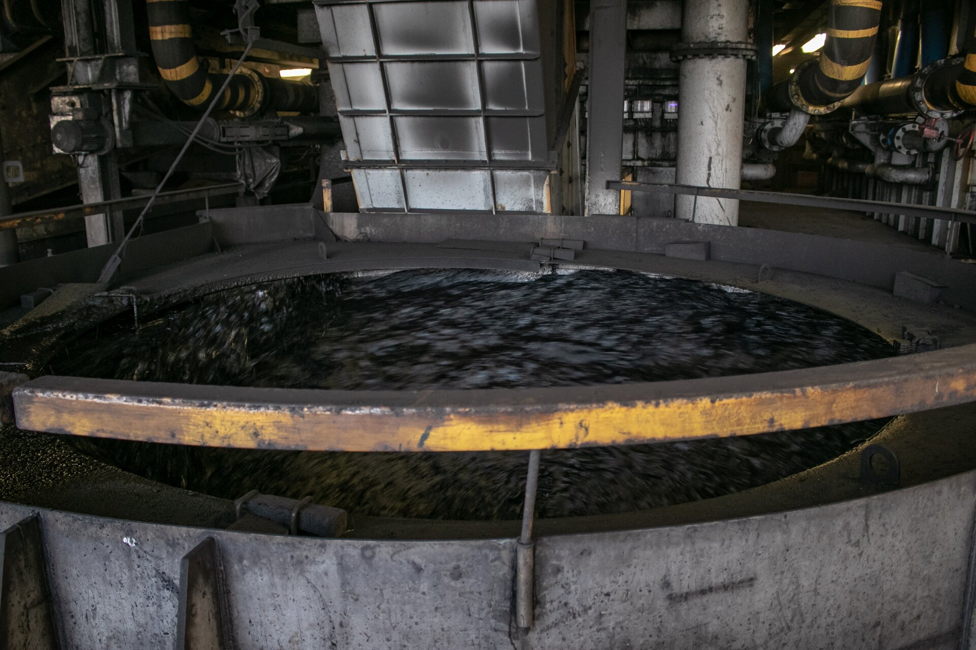 Na zdjęciu znajduje się wirówka sitowa. Wirówka sitowa jest to urządzenie pozwalające na odwadnianie produktów wzbogacania węgla kamiennego.W dolnej części zdjęcia znajduje się cylindryczny pojemnik wirówki. Pojemnik wypełniony jest czarną cieczą. Ciecz jest wprawiona w ruch turbulentny. Za zbiornikiem biegną rury.