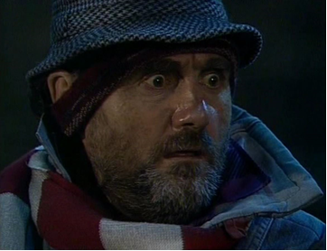 Zdjęcie przedstawia twarz dojrzałego mężczyzny. Jest ona owalna. Mężczyzna ma krótki, szpakowaty zarost. Mężczyzna ma wytrzeszczone oczy. Na głowie ma kapelusz, a pod nim opaskę na uszach. Ubrany jest w kurtkę, pod szyją ma zawinięty szal.