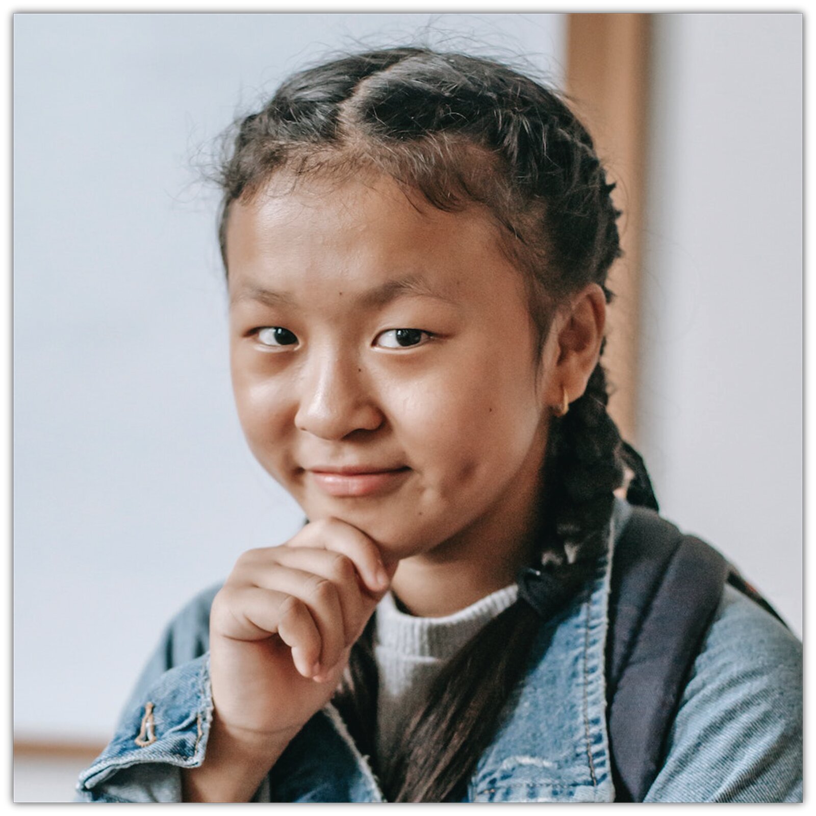 Zdjęcie przedstawia młodą dziewczynę w warkoczykach. Dziewczynka ma na sobie dżinsową kurtkę i na ramieniu niesie plecak. Ręką podpiera podbródek i ma wesoły wyraz twarzy. 