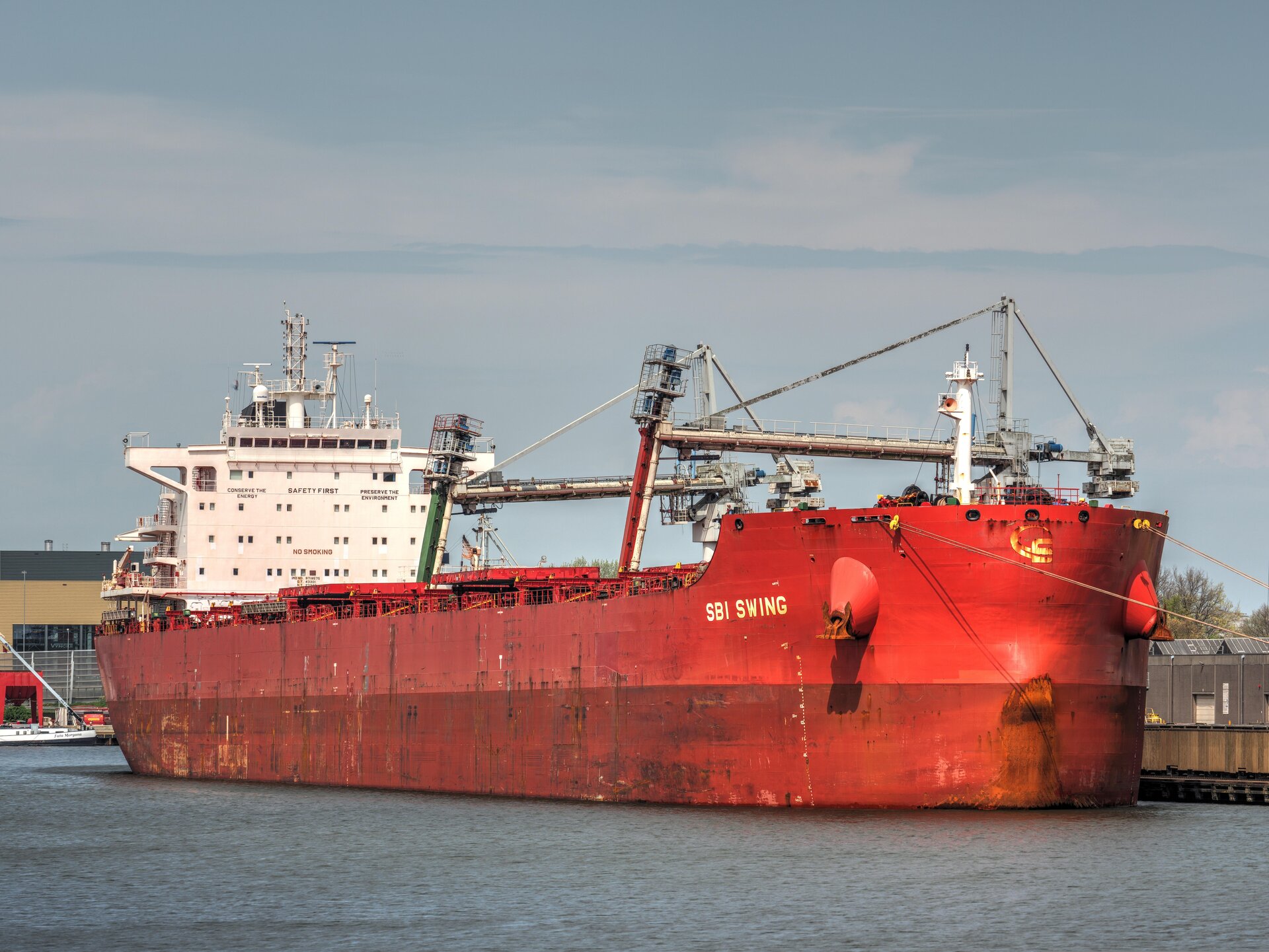 Zdjęcie przedstawia statek tankowiec. Stoi on zacumowany w porcie, z góry do zbiorników podłączone są rury do ich napełniania.