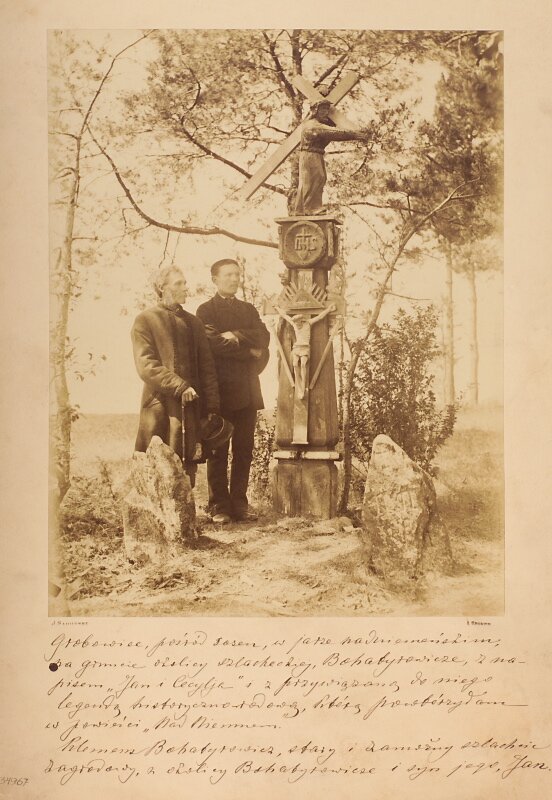 Fotografia przedstawia dwóch mężczyzn stojących na skraju lasu. Jeden z nich jest dojrzały, drugi w sile wieku. Obok nich stoi nagrobek w kształcie ściętego ostrosłupa. Na jego szczycie stoi rzeźba wyobrażająca Chrystusa dźwigającego krzyż. Pod rzeźbą znajduje się monogram IHS. Niżej umieszczono płaskorzeźbę przedstawiającą ukrzyżowanego Chrystusa. Obok grobu stoją dwa duże kamienie. Wokół rosną sosny. Pod ilustracją odręczny napis.