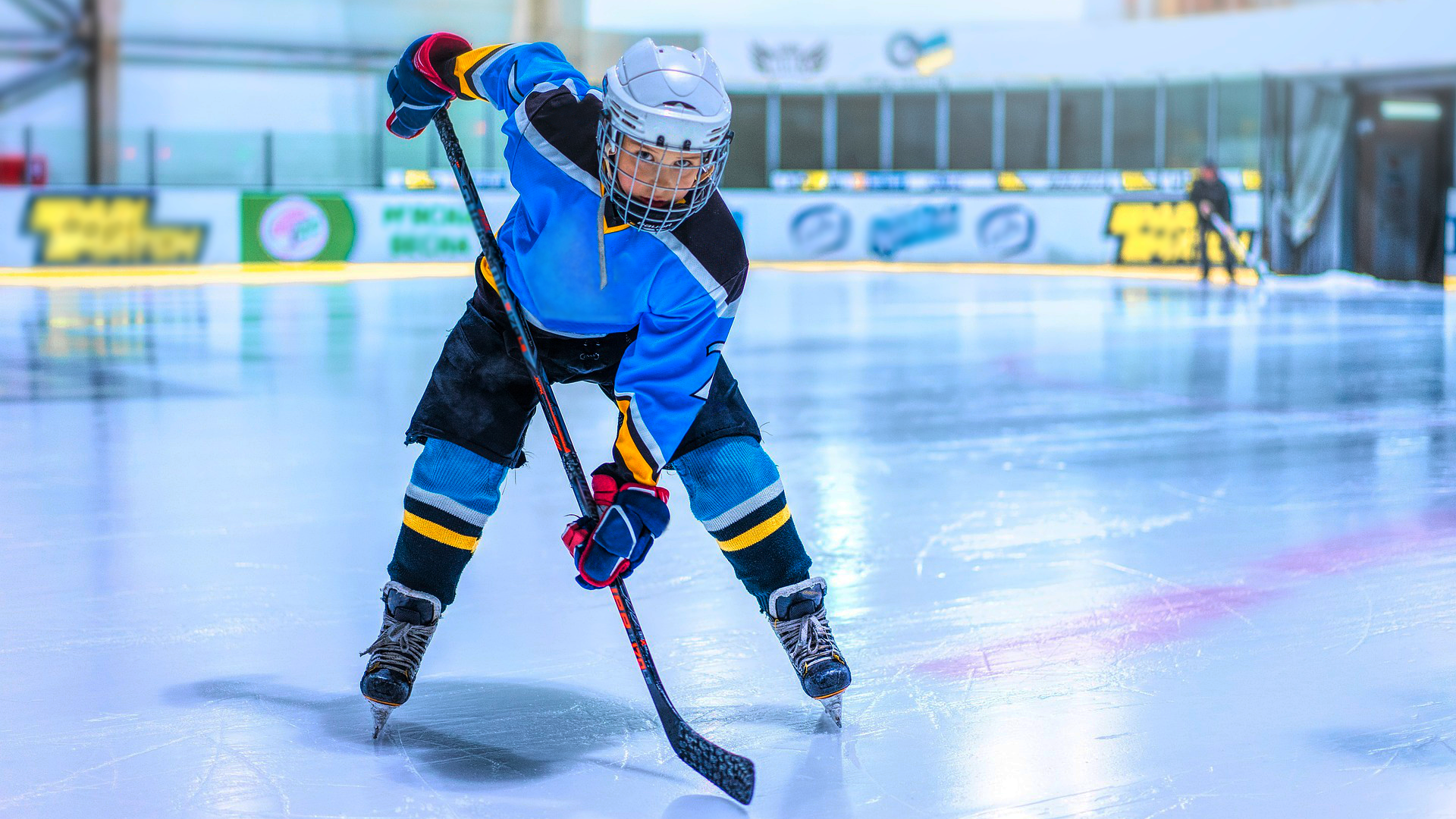 Zdjęcie przedstawia lodowisko, na którym znajduje się hokeista na łyżwach, w kasku z ochronną kratką zakrywającą twarz. Mężczyzna ma na rękach bardzo grube rękawice i trzyma kij hokejowy.