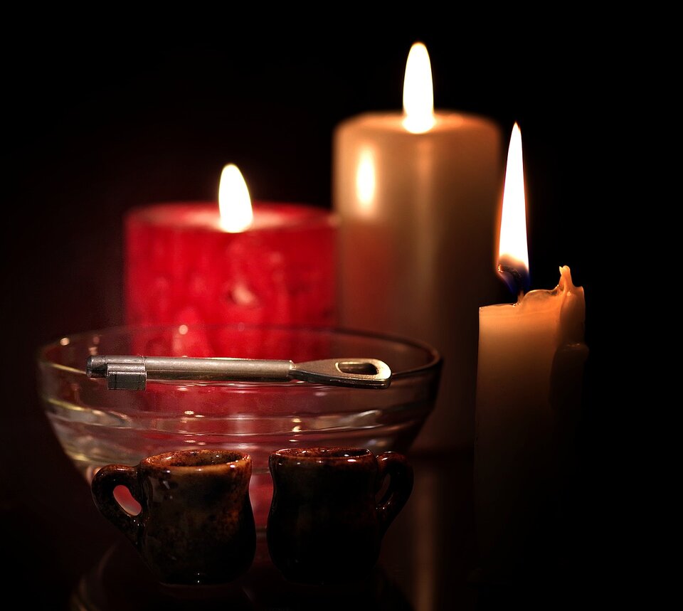 Na zdjęciu, na pierwszym planie znajdują się dwa małe gliniane kubeczki, a za nimi stoi przezroczysta, szklana miska, na której brzegu leży metalowy klucz. W tle znajdują się trzy stojące obok siebie świeczki: czerwona i dwie kremowe. Wszystkie świeczki są zapalone i rzucają ciepłe, przyjemne światło na obiekty umieszczone z przodu ilustracji.