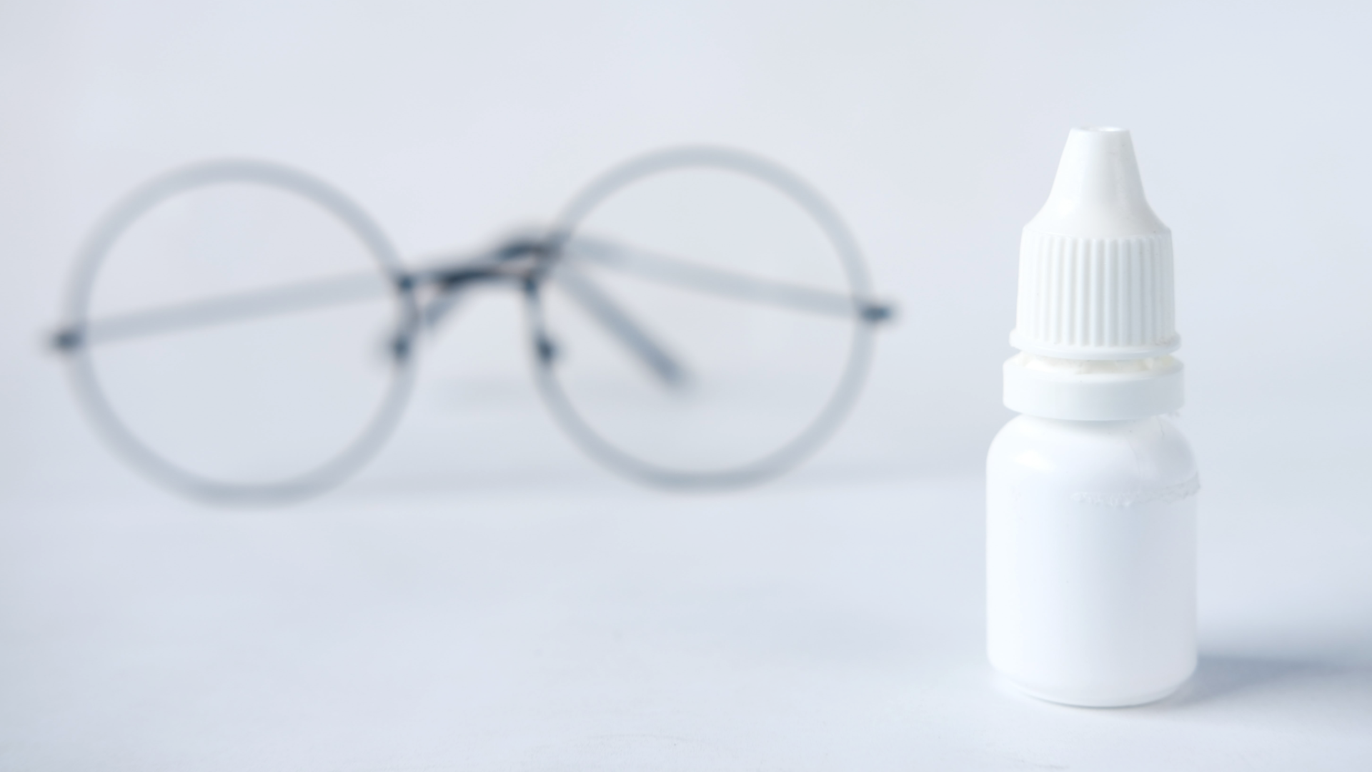 Zdjęcie przedstawia plastikową buteleczkę na krople. W tle widoczne są okulary.