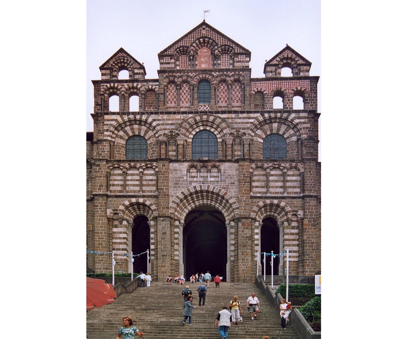 Fotografia barwna przedstawiająca Katedrę Le Puy-en-Velay, Owernia, w 1. poł. XII wieku. Katedra na zewnątrz ma widoczne trzy wieże. Jedna środkowa większa, poboczne mniejsze z futrynami okiennymi. Elewacja jest z czerwonobrunatnej cegły. 