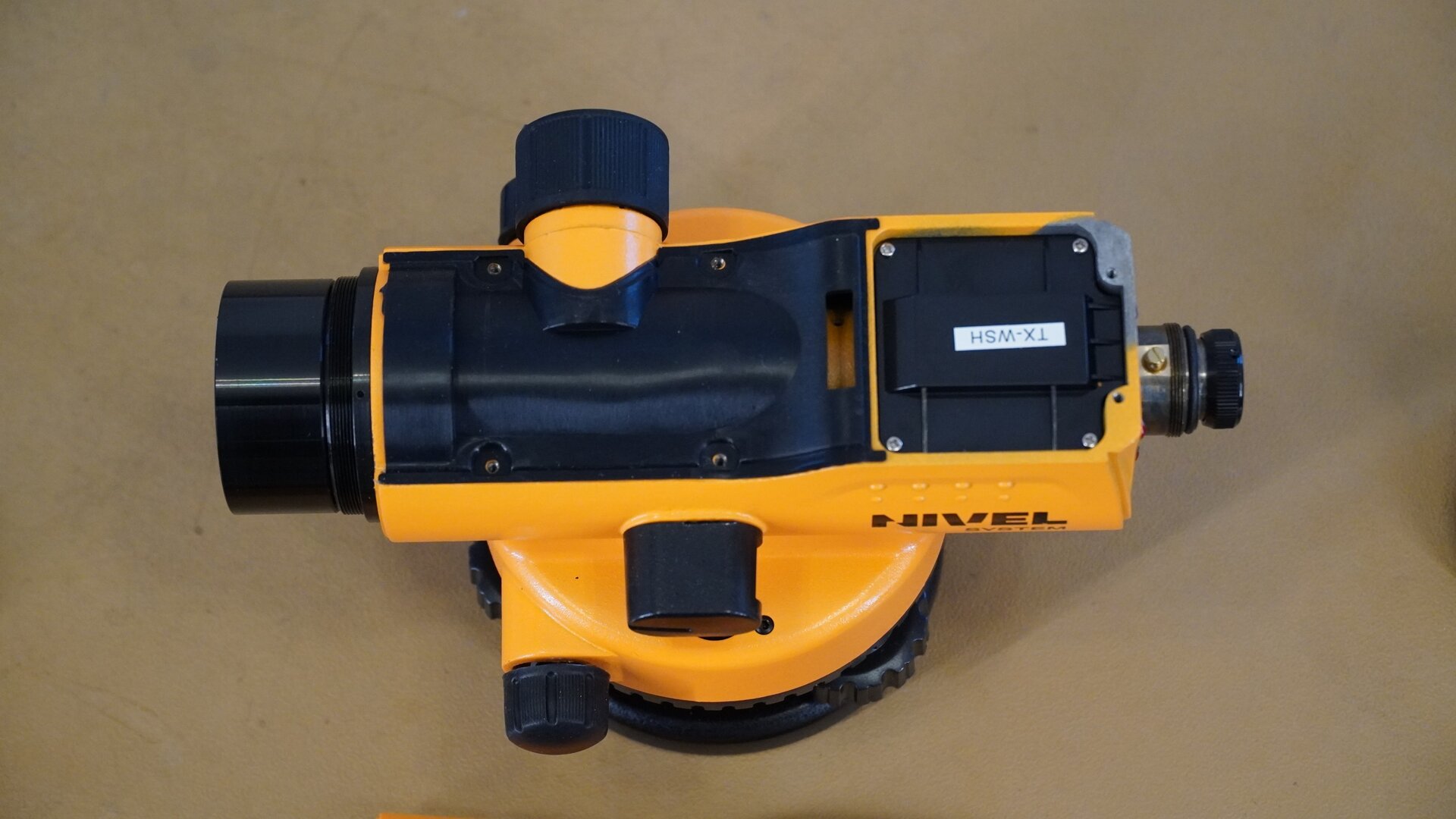 Zdjęcie przedstawia urządzenie widziane od góry. Zdemontowano z niego górną osłonę oraz obudowę okularu. Widoczne są śruby mocujące pokrywę oraz zespół okularu z krzyżem nitek. Pierścień okularu ma postać czarnego pokrętła, które reguluje ostrość krzyża nitek. Tuż za nim zabudowany jest kompensator.