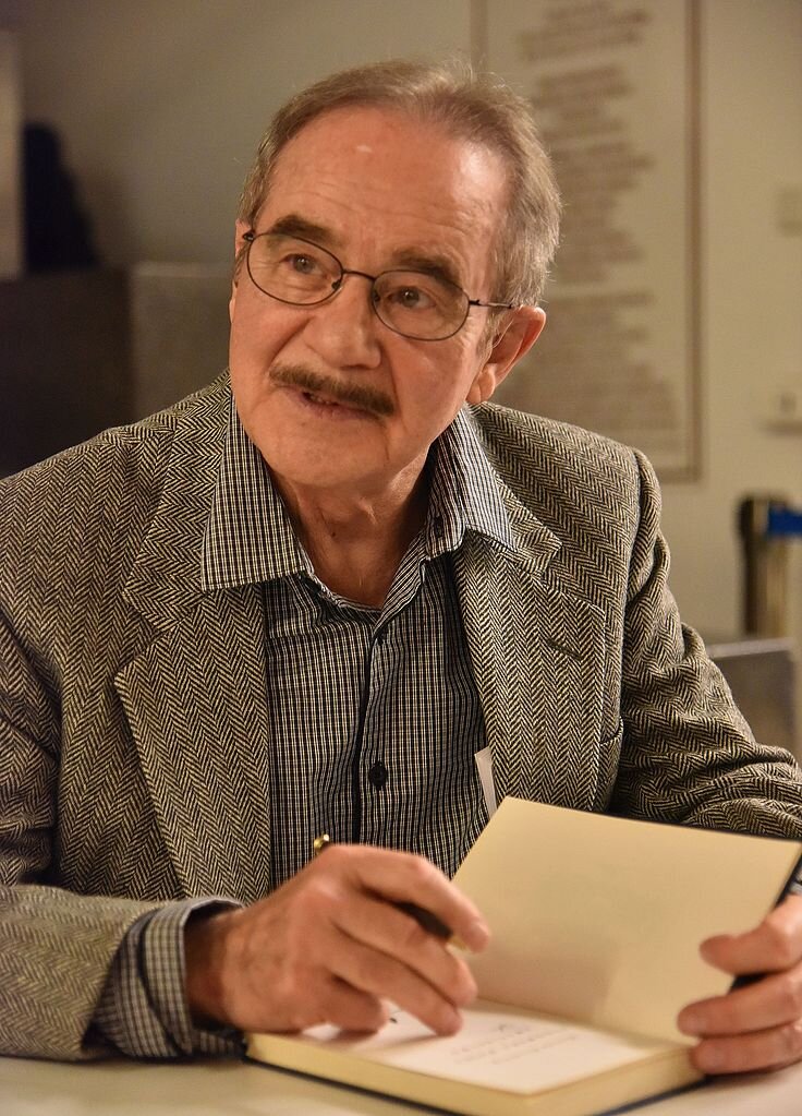 Zdjęcie przedstawia starszego mężczyznę od pasa w górę. Jego twarz jest owalna, są na niej liczne zmarszczki. Mężczyzna ma szeroki noc, wysokie czoło, nosi okulary. Jego włosy są rzadkie, krótko ostrzyżone. Mężczyzna ubrany jest w marynarkę i koszulę. Mężczyzna siedzi przy stole. Trzyma lewą dłonią otwartą książkę. W prawej dłoni ma długopis.