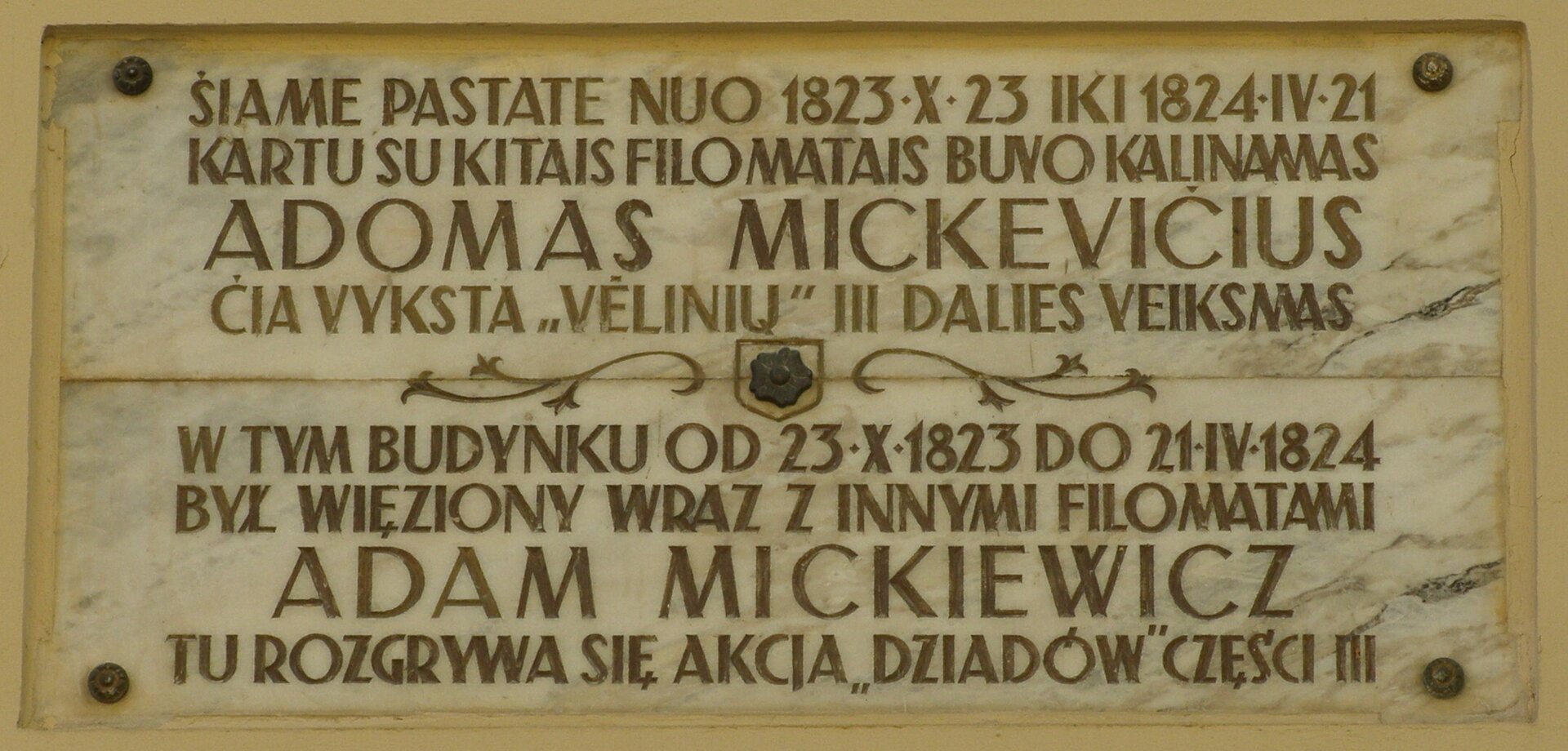 Zdjęcie przedstawia tablicę pamiątkową. Widnieje na niej napis w języku litewskim i polskim: W tym budynku od 23 października 1823 roku do 21 kwietnia 1824 roku był więziony wraz z innymi filomatami Adam Mickiewicz. Tu rozgrywa się akcja Dziadów części III.