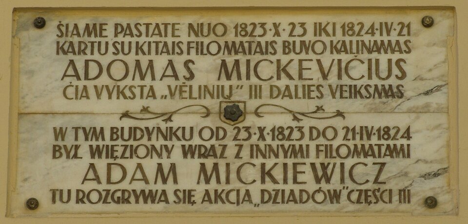 Zdjęcie przedstawia tablicę pamiątkową. Widnieje na niej napis w języku litewskim i polskim: W tym budynku od 23 października 1823 roku do 21 kwietnia 1824 roku był więziony wraz z innymi filomatami Adam Mickiewicz. Tu rozgrywa się akcja Dziadów części III.