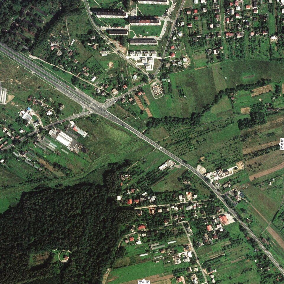 Zdjęcie przedstawia węzeł Wielicka autostrady A4 w 1996 roku. Przedstawiona jest na nim dwupasmowa droga, która od momentu skrzyżowania rozdziela się na dwa dwupasmowe odcinki, oddzielone pasem zieleni. Skrzyżowanie przecina drogę, ta część ulicy prowadzi przez pola i tereny zamieszkałe, na górze zdjęcia rozgałęzia się w dwóch kierunkach. Druga strona przecinającej autostradę ulicy również prowadzi do budynków. Na dole zdjęcia znajduje się las oraz uchwycone z góry osiedle domków jednorodzinnych.