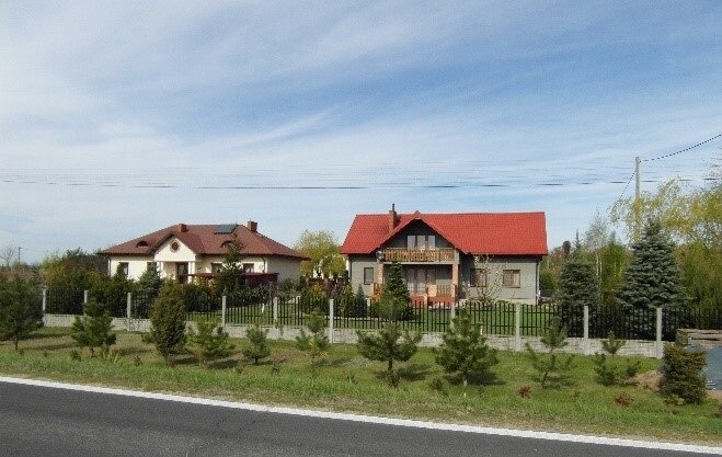 Zdjęcie przedstawia domy mieszkalne. Z prawej strony dom z czerwonym dachem i szarą elewacją ogrodzony płotem, po lewej biały budynek z bordowym dachem. Przed domami metalowe ogrodzenia z betonowymi słupkami.