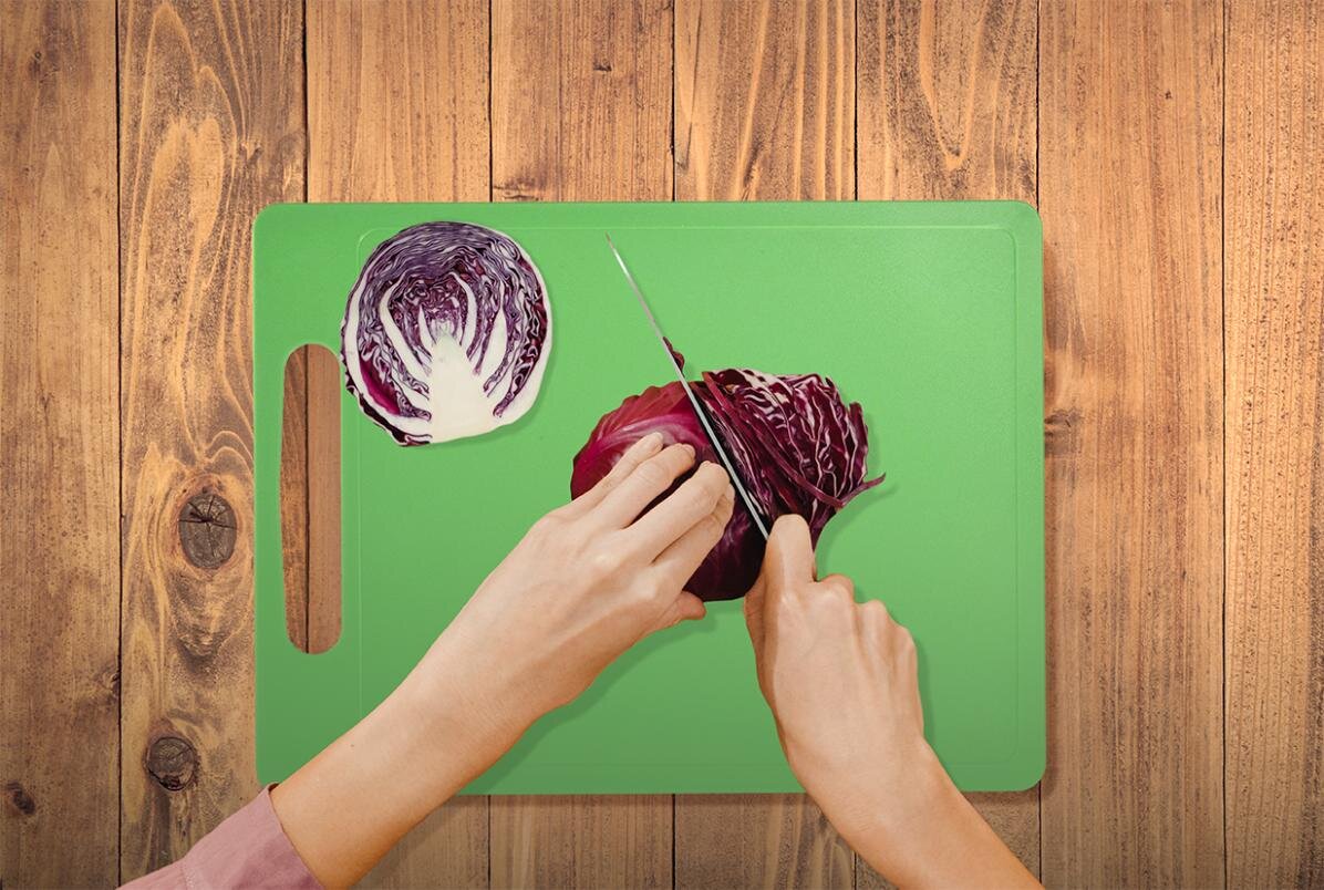 Zdjęcie przedstawia technikę szatkowania kapusty. Pośrodku widoczna jest zielona deska, na której znajduje się połowa główki czerwonej kapusty, oraz połówka główki przytrzymywana lewą ręką szatkowana na paski. W prawej ręce widoczny jest nóż szatkujący kapustę.
