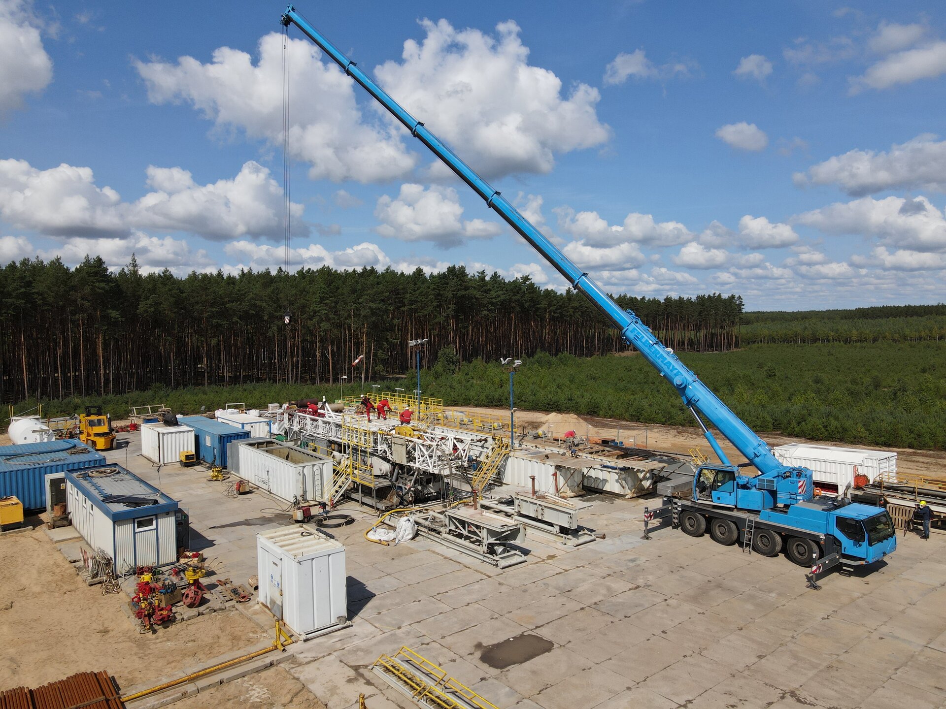 Na zdjęciu widoczny jest proces podnoszenia masztu wiertniczego. Po prawej stronie zdjęcia widać niebieski podnośnik znajdujący się na podwoziu samochodowym. Podnośnik znajduje się na terenie betonowego placu wiertniczego. Ma opuszczoną linę do której robotnicy przywiązują stalową konstrukcję kratową. Konstrukcja leży na rozłożonym podeście. Po lewej stronie podnoszonego masztu znajduje się biały prostopadłościenny zbiornik przypominający kontener. Na terenie całego placu ustawione są białe i niebieskie prostopadłościany przypominające kontenery.W tle zdjęcia widać drzewa.