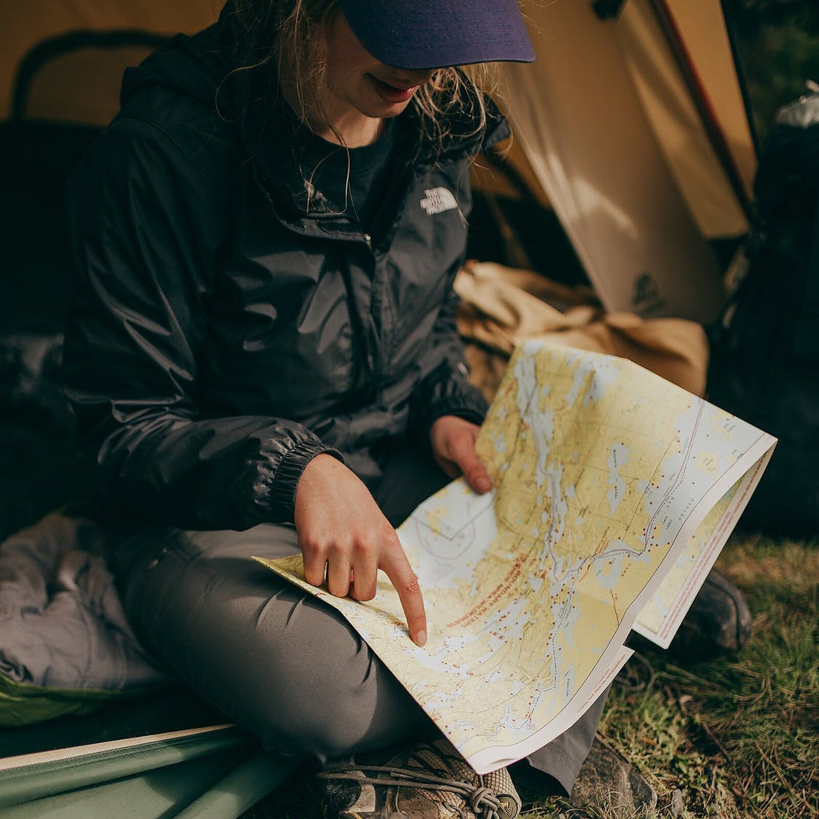 Zdjęcie przedstawia kobietę siedzącą ze skrzyżowanymi nogami, na progu namiotu. Kobieta trzyma w rękach mapę, opierając ją na nogach. Ma na sobie czapkę z daszkiem, przeciwdeszczową kurtkę oraz długie spodnie.