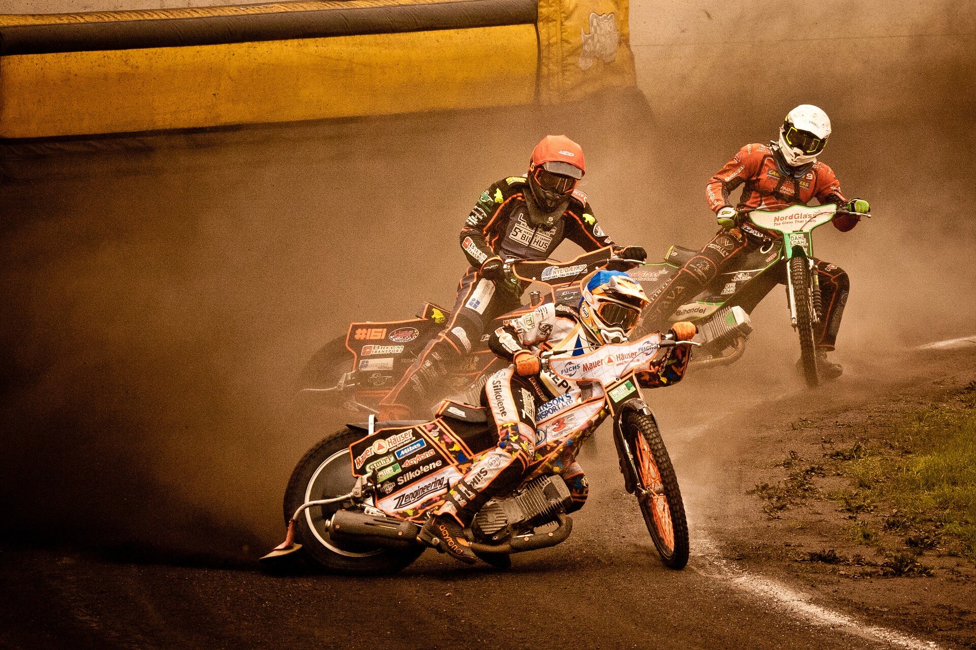 Zdjęcie przedstawia tor żużlowy w trakcie wyścigu. Na pierwszym planie można dostrzec trzech zawodników, którzy jadą na sportowych motocyklach o lekkiej konstrukcji. Zawodnicy ubrani są w kolorowe kombinezony, które są wyposażone w ochraniacze na nogach i rękach. Na ich głowach widnieją kaski oraz okulary ochronne, które chronią ich przed kurzem.
