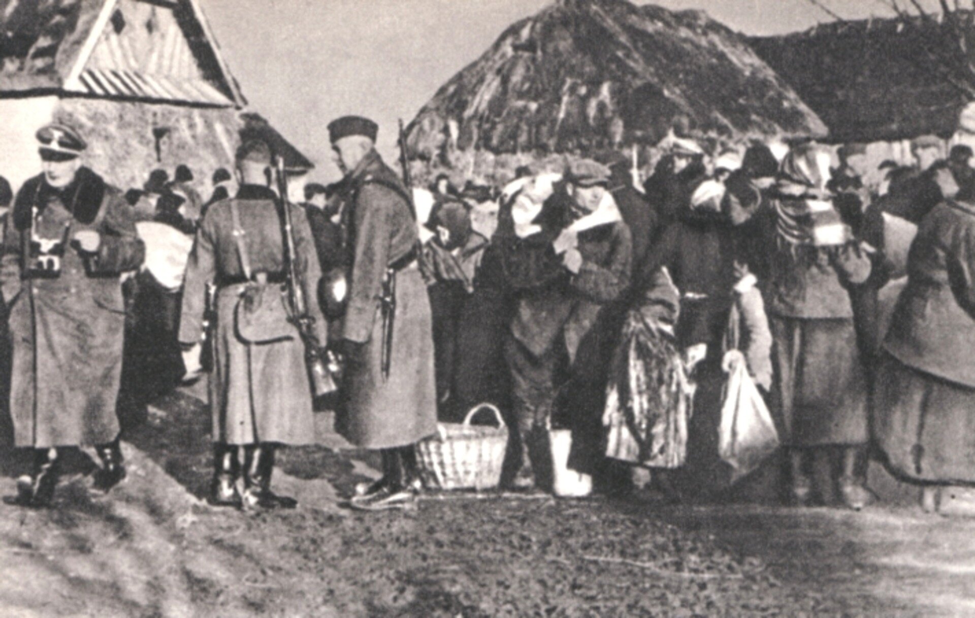 Fotografia czarno-biała przedstawiająca polskich rolników wysiedlonych z farm przez SS. Widoczny jest tłum ludzi, który wygląda, jakby miał zaraz rozpocząć wędówkę. Niemal każda osoba ma ze sobą torbę lub kosz. W tle widoczne są dachy wiejskich domostw.