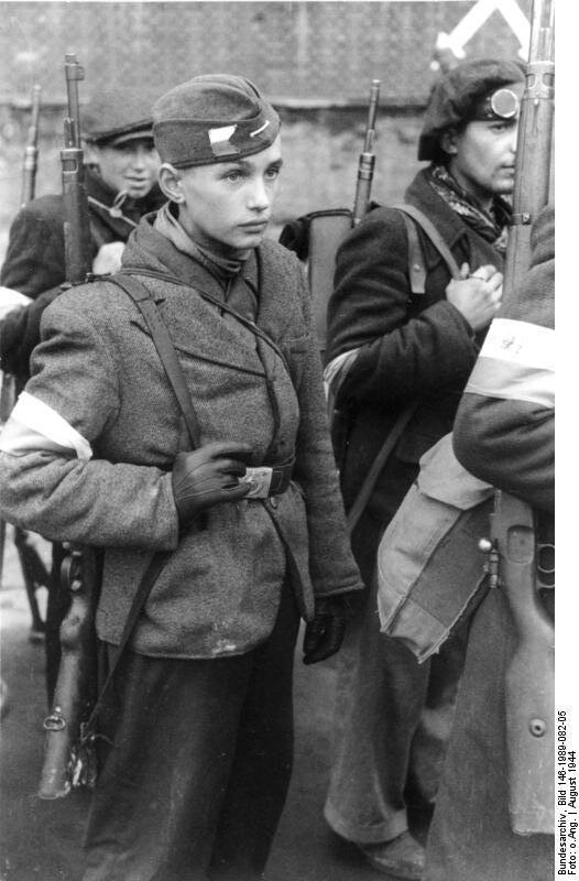 Kliknij, aby powiększyć. Ilustracja przedstawia część zgrupowania - dokładnie trzech młodych powstańców. Stoją w szeregu. Mają przełożoną przez ramię broń. Opaski w barwach narodowych na ramieniu. Są bardzo młodzi. Chłopiec na pierwszym planie nosi cywilną marynarkę. Na czapce ma barwy polskie narodowe.