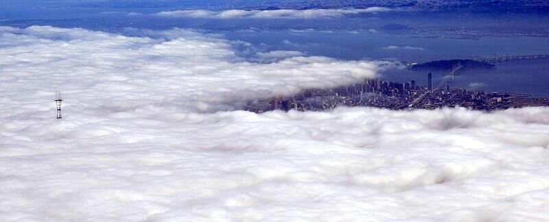 Zdjęcie przedstawia warstwę białych chmur wiszących nisko nad miastem. Po prawej stronie spod warstwy chmur wyłaniają się budynki miasta oraz ocean.  