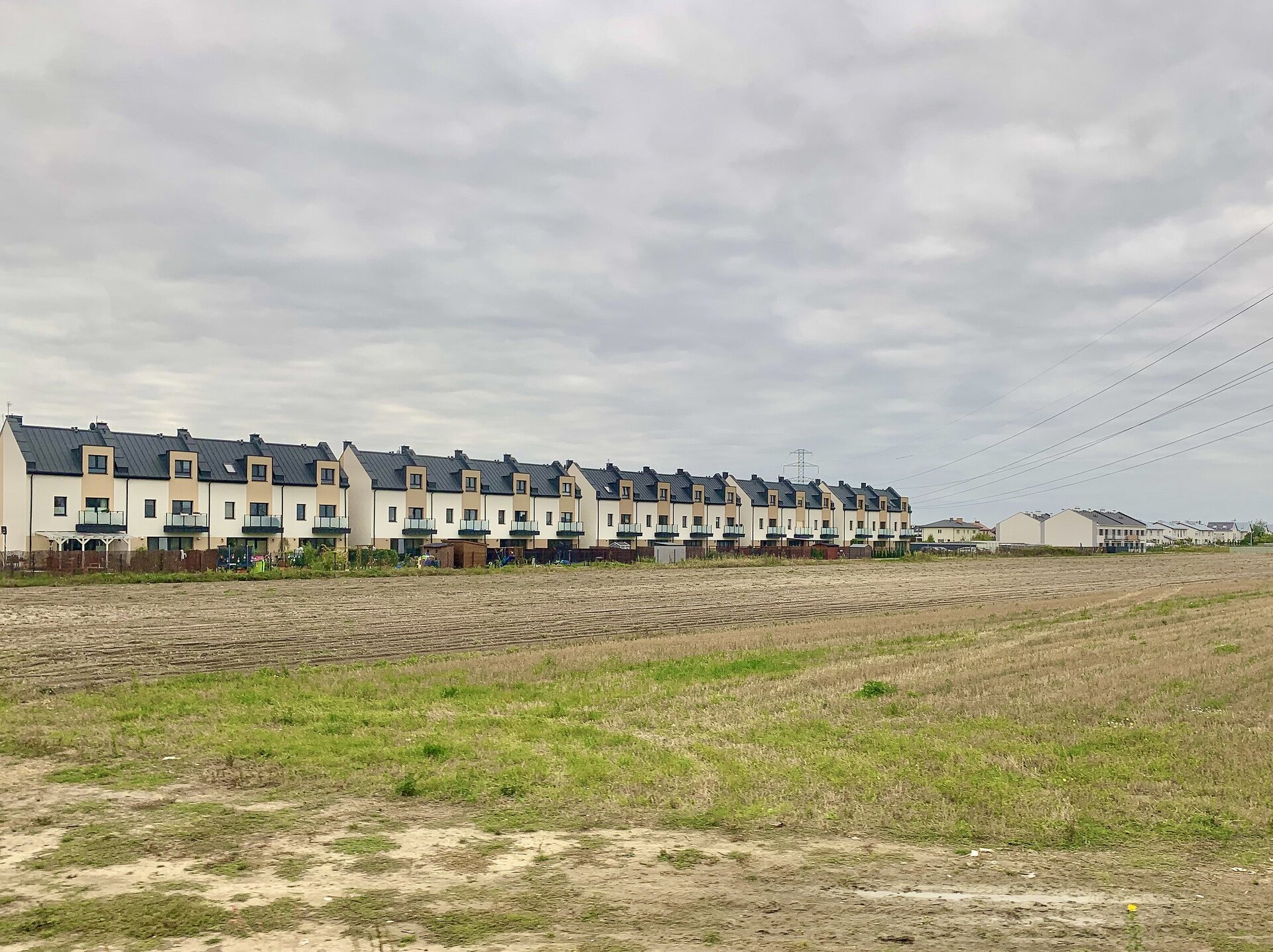 Fotografia przedstawiająca domy szeregowe na terenie podmiejskiej wsi. Zwarta zabudowa, domy stoją blisko siebie wzdłuż ulicy. Jeden budynek tworzą połączone cztery identyczne segmenty, na zdjęciu cztery budynki. Na drugim planie podobna zabudowa, na pierwszym planie, tuż przed budynkami, pole uprawne.