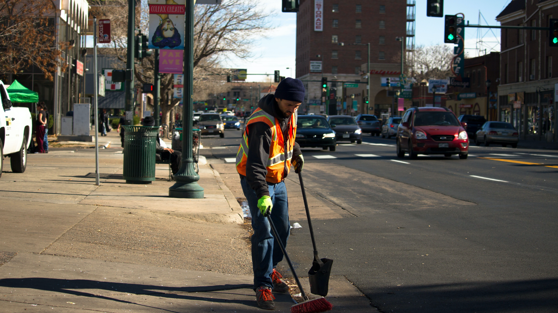 Zdjęcie przedstawia mężczyznę sprzątającego ulicę. Mężczyzna ubrany jest w kamizelkę odblaskową. W rękach trzyma szufelkę i szczotkę. W tle widoczne są samochody jadące ulicą, sygnalizację świetlną, chodnik, budynki.