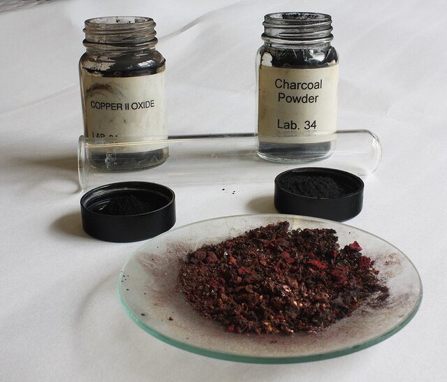 Ilustracja przedstawia czarno-czerwoną substancję sproszkowaną na szklanym spodku.  Za spodkiem stoją dwa szklane słoiczki zawierające etykiety z anglojęzycznymi napisami: copper 2 oxide i charcoal powder.