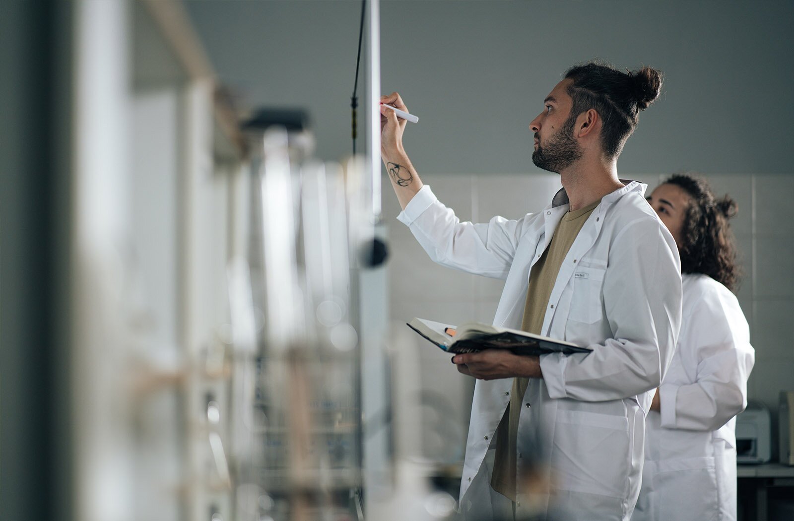 Na zdjęciu widoczne są dwie osoby: młody mężczyzna i młoda kobieta. Obydwoje są ubrani w białe fartuchy i stoją przed tablicą. Mężczyzna prawą ręką uniesioną do góry zapisuje coś na tablicy, zaś w lewej ręce trzyma otwarty notes. Kobieta stoi za nim, ma lekko uniesioną do góry głowę i patrzy na tablicę.