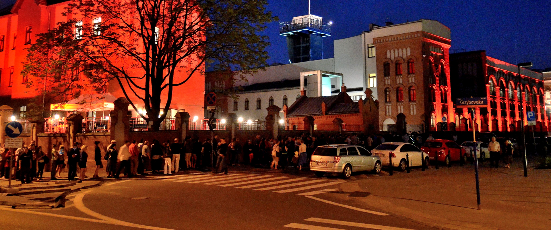 Zdjęcie przedstawia ludzi oczekujących w kolejce na wejście do muzeum w noc muzeów. Na zdjęciu widoczne są oświetlone budynki oraz zaparkowane samochody.