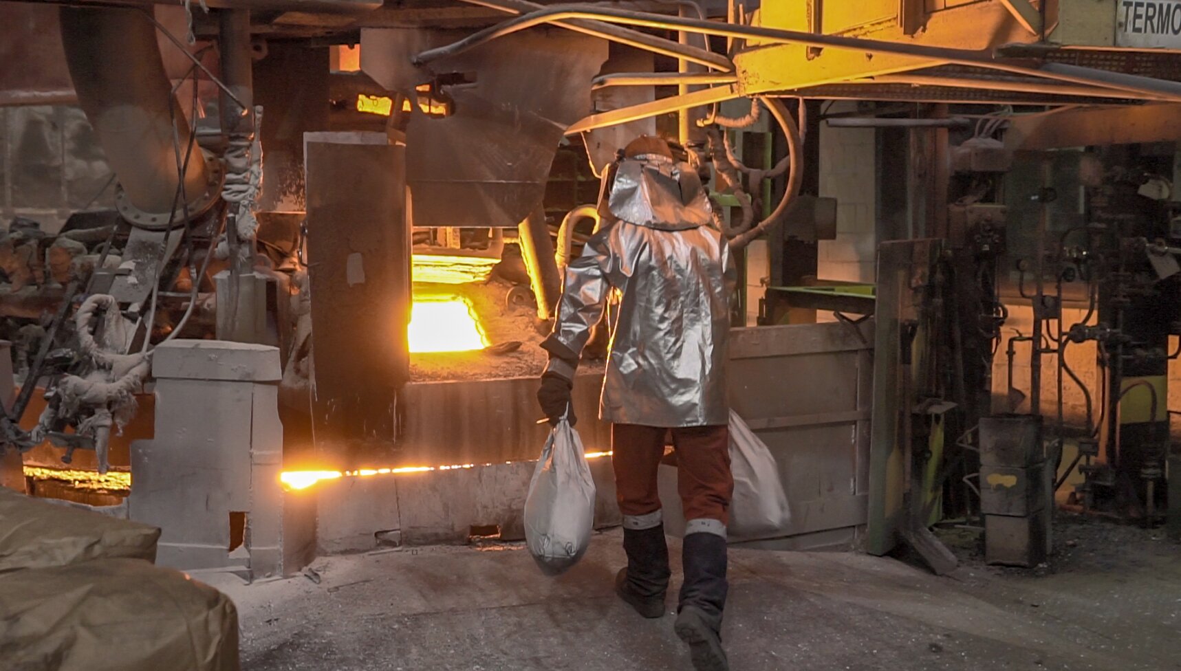 Na zdjęciu widać proces dozowania oknem roboczym. W centralnej części zdjęcia widać piec. W przedniej ścianie znajduje się okno robocze. Okno ma kształt prostokątny. Przez okno widać płomienie. Do pieca podchodzi pracownik ubrany w srebrny strój ognioodpornym. W rękach trzyma dwa białe worki.