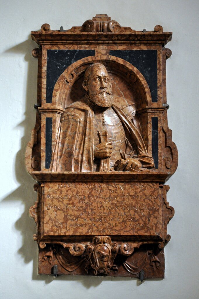 Zdjęcie przedstawia tablicę pamiątkową poświęconą Janowi Kochanowskiemu. Jest to przyścienna tablica z wyrzeźbionym popiersiem wieszcza umieszczonym w dekoracyjnej arkadzie oraz tekstem pochwalnym poniżej. Wykonana jest z dwóch gatunków marmuru. Elementy płaskorzeźbione nawiązują do architektury renesansowej.