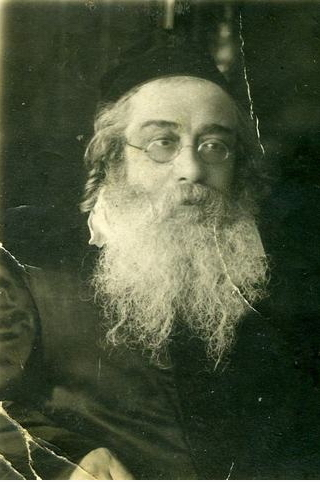 Czarno‑białe zdjęcie przedstawia starszego mężczyznę. Ma on długą siwą brodę, okulary, a na głowie czarną jarmułkę. Jest ubrany w czarny strój. 