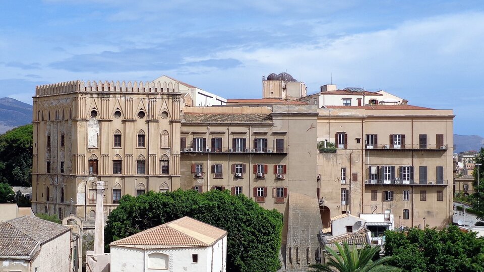 Zdjęcie przedstawia budynek kilkupiętrowego, dużego pałacu. Składa się z rzędów okien oraz brązowej elewacji. Okna na prawej części budynku mają drewniane okiennice. Lewa strona budynku składa się z płaskich kolumn, dekoracji w kształcie ostrych łuków oraz rzędów okien. Wokół stoją niższe budynki. W tle zalesione wzgórza i niebo z chmurami.