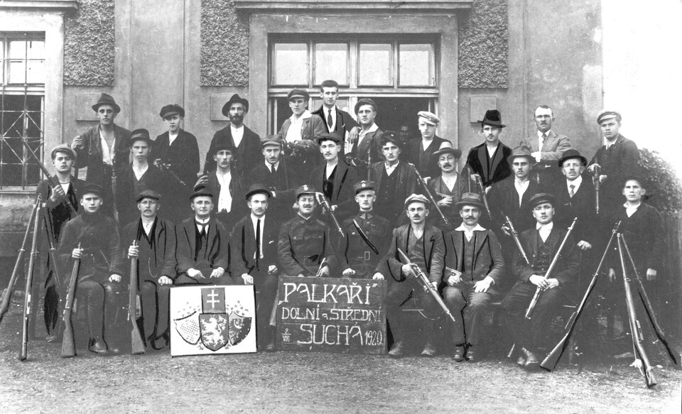 Zdjęcie przedstawia żołnierzy z bronią, którzy siedzą i stoją w kilku rzędach. Są różnie ubrani: w garnitury, mundury, czapki, kapelusze oraz kaszkiety. Są to głównie młodzi mężczyźni. W pierwszym rzędzie widoczne są dwie tabliczki: jedna z godłami, a druga z napisem w innym języku z rokiem 1920. Za mężczyznami widoczna jest ściana budynku z oknami.