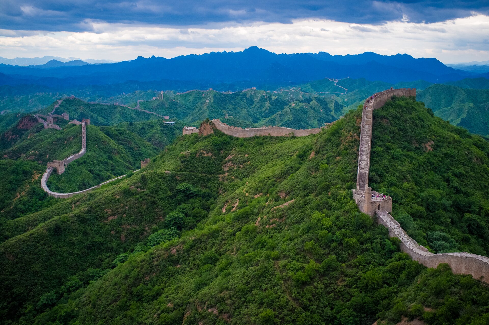 Na zdjęciu Wielki Mur Chiński, przebiegający po szczytowych partiach pasma górskiego porośniętego lasami, wieże sygnalizacyjne. Na murze turyści.
