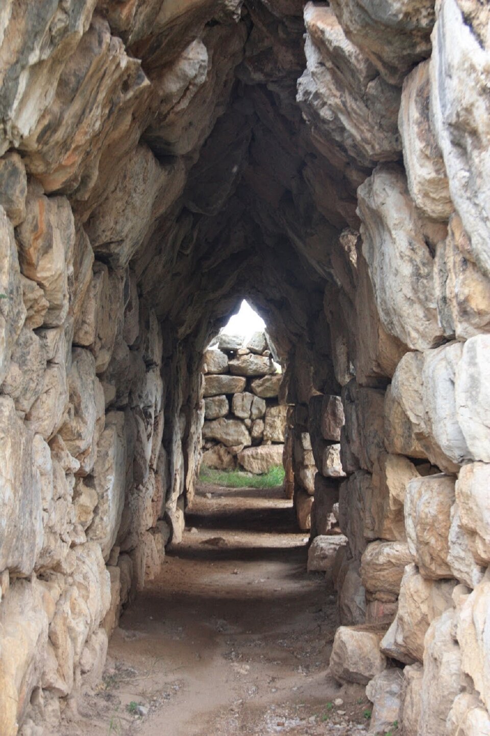 Fotografia przedstawia fragment potężnych murów obronnych w Tirynsie na Peloponezie. Ukazuje masywną konstrukcję zbudowaną z ogromnych nieobrobionych bloków kamiennych.  Mur ma     nieregularną fakturę, kamienie są różnej wielkości i kształtu. Budowla jest masywna. Na dalszym planie widoczna jest różnica poziomów, co wskazuje na wzniesienie terenu, na którym zbudowano twierdzę.
