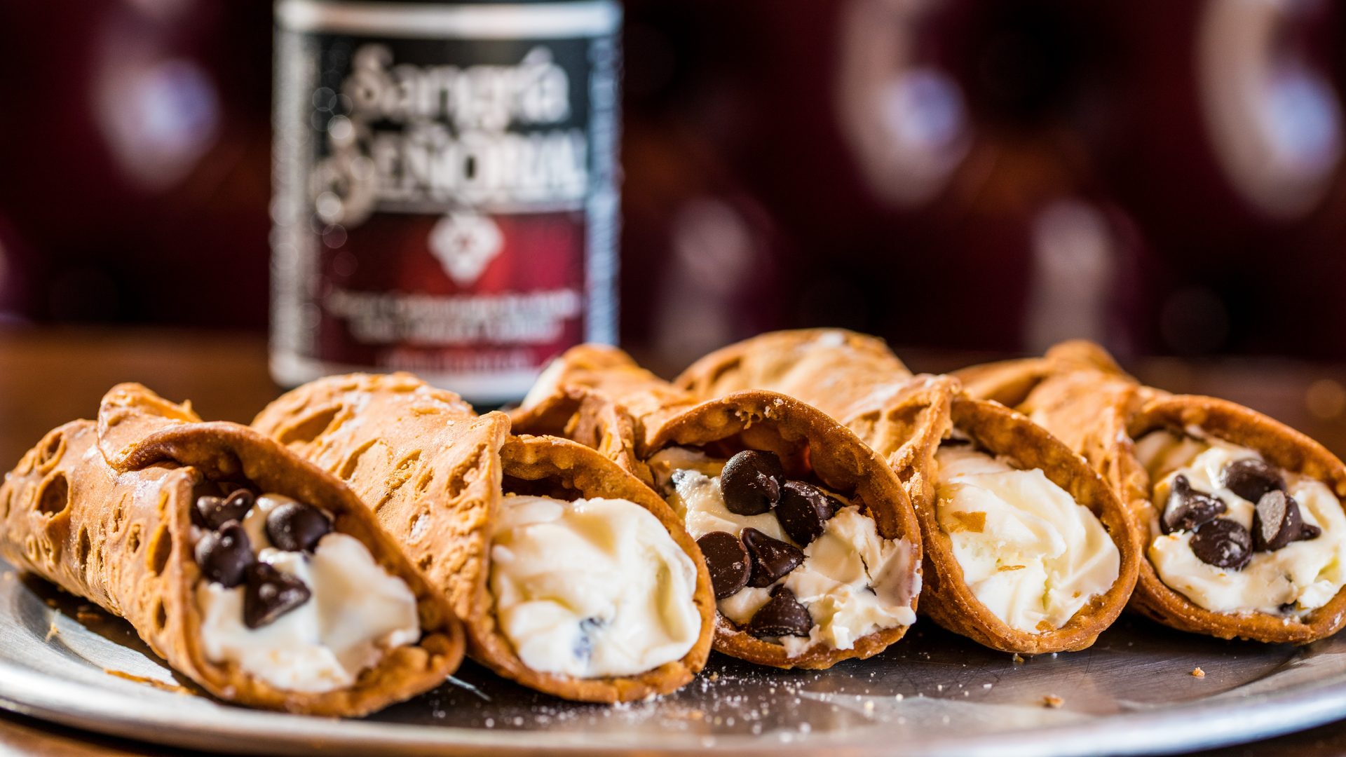 Na zdjęciu znajduje się pięć Cannoli siciliani, które leżą na metalowej tacy. Każdy wypełniony jest kremem, a niektóre dodatkowo kawałkami czekolady.