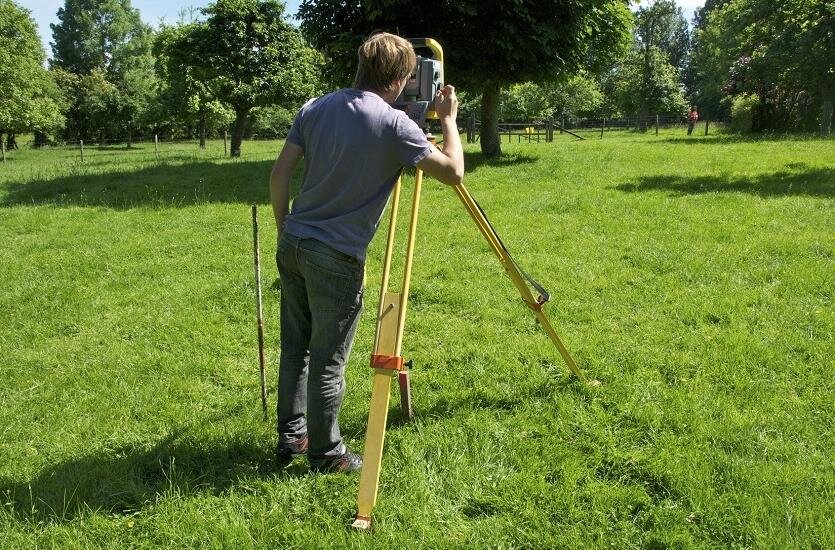 Fotografia przedstawia geodetę podczas wyznaczania punktów granicznych. Czynność ta odbywa się za pomocą profesjonalnego sprzętu pomiarowego, dzięki któremu geodeta może sprawdzić, czy dany punkt geodezyjny uległ przesunięciu lub czy został zniszczony.