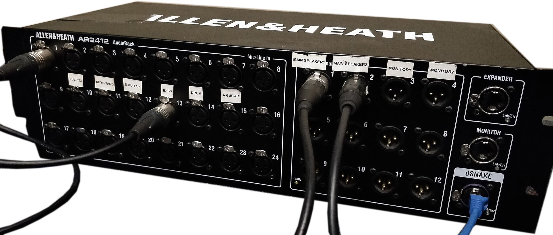 Zdjęcie przedstawia stagebox. Widoczny jest czarny prostopadłościan, w ujęciu trzy czwarte. Na przedniej ścianie prostopadłościanu widoczne są liczne kółka – gniazda do wtyczek. Widoczne są cztery wtyczki z czarnymi kablami wpięte w gniazda. W prawym dolnym rogu widoczny jest niebieski kabel wpięty w gniazdo. Na górnej ścianie prostopadłościanu widoczny jest napis z dużych białych liter – nazwa firmy.