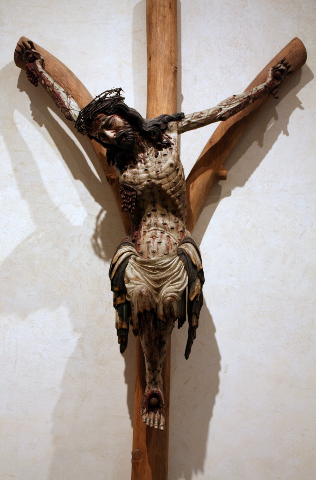 Ilustracja przedstawia figurę, na której widoczny jest Jezus wiszący na krzyżu z licznymi ranami na ciele. Rzeźba wykonana jest z drewna.