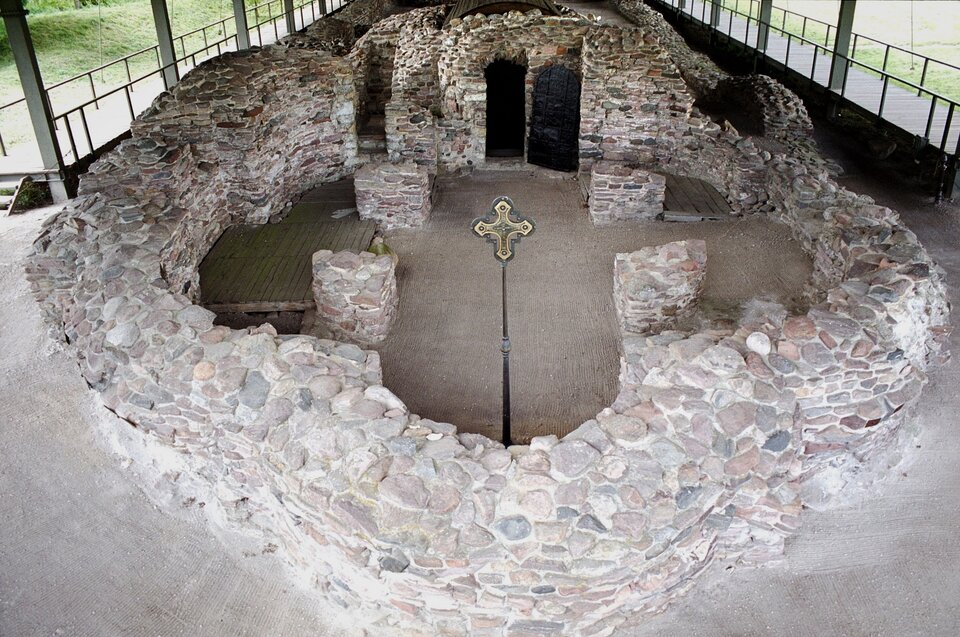 Ilustracja o kształcie poziomego prostokąta przedstawia fotografię palatium położonego na największej wyspie jeziora Lednicy (Ostrów Lednicki). Na wykonanym z górnego widoku zdjęciu widoczne są pozostałości po palatium (przypałacowej kaplicy). Fragmenty murów zbudowanych z kamienia uformowane są w kształt krzyża greckiego. Za murami znajduje się ogrodzenie.