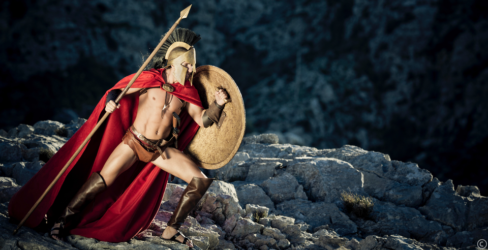 Zdjęcie uwspółcześnione. Mężczyzna ma na biodrach przepaskę z pasem, na łydkach nagolenniki ze skóry, na stopach sandały, na ramionach czerwony płaszcz, w lewej ręce trzyma metalową, okrągłą tarczę, w prawej ręce trzyma drewnianą włócznię z metalowym ostrzem. Na głowie ma hełm z grzebieniem. Przedstawiony jest z lewej strony zdjęcia, pochylony ciałem ku prawej krawędzi, opiera ciężar na lewej nodze wysuniętej do przodu, prawa cofnięta z tyłu. W tle wzgórza i wzniesienie kamienne.