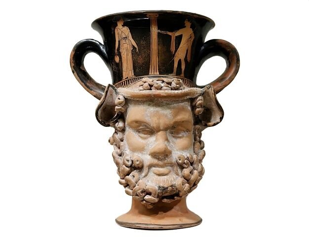 Na zdjęciu przedstawiono naczynie. Jego podstawą jest głowa brodatego mężczyzny. Na głowie mężczyzny znajduje się waza z dwoma uchwytami. Na wazie przedstawiono malowidła dwie postaci stojące po dwóch stronach kolumny. Postać po lewej stronie ubrana jest w długą suknię, postać po prawej, to nagi mężczyzna, z płaszczem przerzuconym przez ramiona.