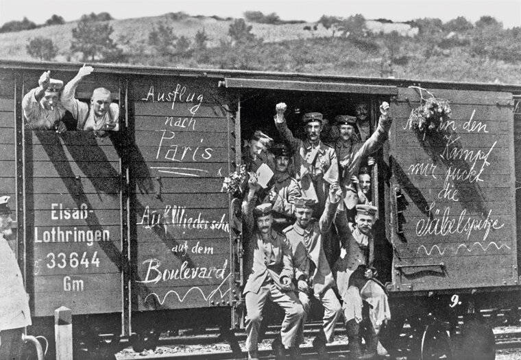 Zdjęcie przedstawia mężczyzn w mundurach, którzy machają radośnie . Stoją i ziedzą  w wejściu do wagonu pociągu. Wagon jest drewniany. Znajdują się na nim namalowane napisy w języku niemieckim.