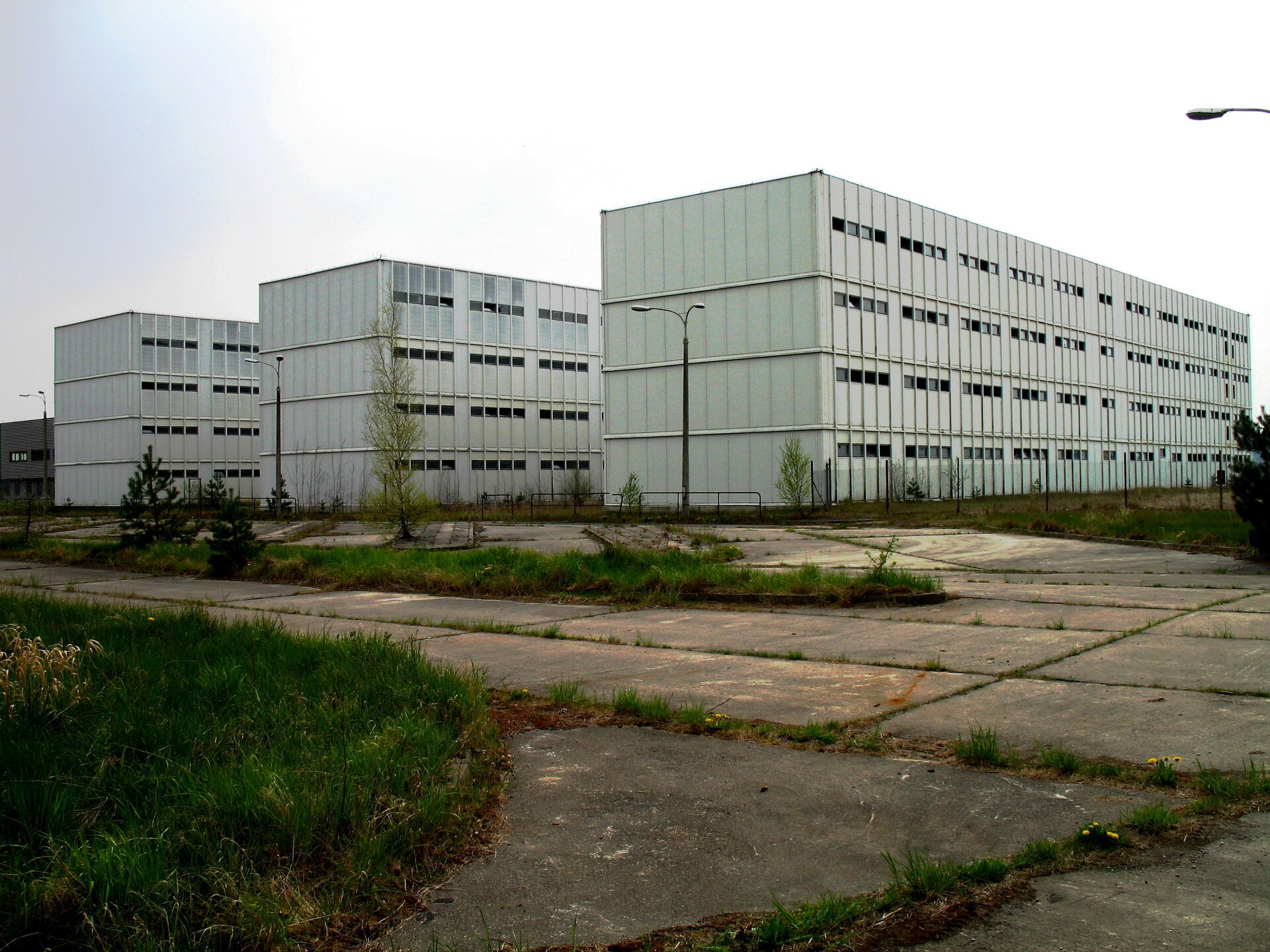 Ilustracja przedstawia niemal całkowicie ukończone budynki dla pracowników. Grafika ukazuję trzy prostokątne bloki koloru białego, postawione równolegle do siebie. W koło budowli rozciąga się teren zarośnięty trawą jak i zalany betonem.