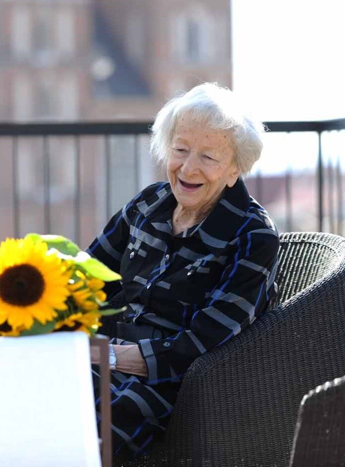 Fotografia przedstawiająca starszą, uśmiechniętą kobietę - Wisławę Szymborską. Ma siwe, krótkie włosy. Jest kobietą o drobnej sylwetce. Ubrana jest w sukienkę. Siedzi w fotelu na tarasie lub balkonie, w dłoniach trzyma bukiet słoneczników.