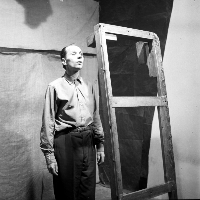 Zdjęcie przedstawia dojrzałego mężczyznę stojącego na baczność. Mężczyzna ubrany jest w koszulę i spodnie. Obok niego stoi w pionie rama drewnianego łóżka. W tle rozwieszone płachty materiału. 