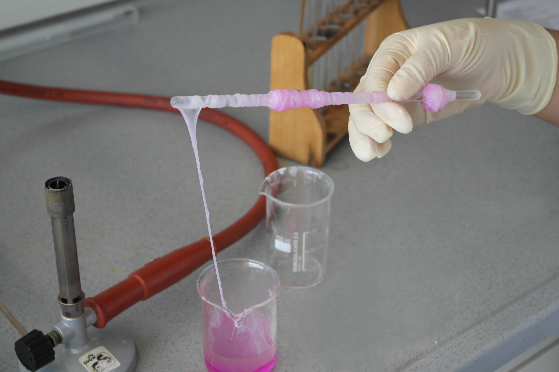 Zdjęcie przedstawia uzyskany nylon. W tle widnieje palnik i drewniany stojak na probówki, natomiast w centrum znajdują się dwie zlewki. W jednej zlewce widoczna jest różowa kleista substancja. Nad zlewką z tą substancją widoczna jest dłoń, która trzyma szklaną bagietkę, na którą nawinięte są ciągnące się fragmenty substancji.