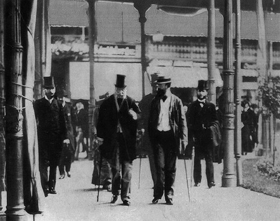 Na czarno‑białym zdjęciu idą dwaj mężczyźni. Są ubrani w koszule i czarne płaszcze. Obaj mają laseczki. Mężczyzna po lewej ma siwą brodę i czarny cylinder, a ten po prawej długa czarną brodę i kapelusz. Mężczyźni dyskutują. Za nimi idą inni gustownie ubrani mężczyźni. W oddali znajdują się budynki. 