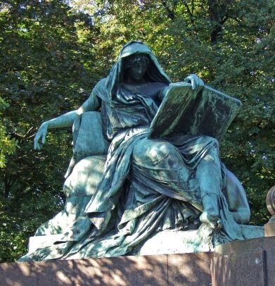 Fotografia przedstawia wyrzeźbioną ludzką postać. Rzeźba jest  w kolorze zielono- czarnym; stoi na brązowej podstawie. Przedstawia siedzącą postać. Kobieta ubrana jest w długie szaty, na głowę ma zarzuconą chustę. W lewej dłoni trzyma otwartą księgę, a prawa spoczywa na oparciu krzesła. Tło stanowią zielone drzewa.