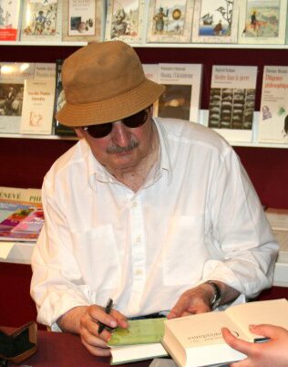 Kliknij, aby powiększyć Zdjęcie mężczyzny w kapeluszu, ciemnych okularach i w białej koszuli. Ma wąs i wydatny nos. W ręce trzyma długopis i książkę. Ktoś spoza kadru podsuwa mu otwartą książkę prawdopodobnie do podpisu. W tle regał z książkami.